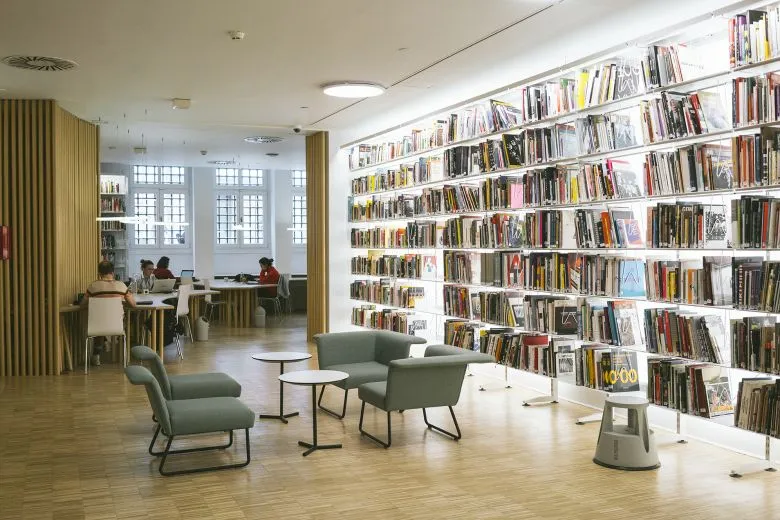 SALT Galata Kütüphanesi'nde, modern tasarımlı aydınlatmalı raflarda sanat ve mimarlık koleksiyonundan yüzlerce kitabın sergilendiği, ön planda konforlu koltukların bulunduğu, arka planda ise araştırmacıların uzun çalışma masalarında odaklanarak çalıştığı huzurlu bir ortam görünüyor.