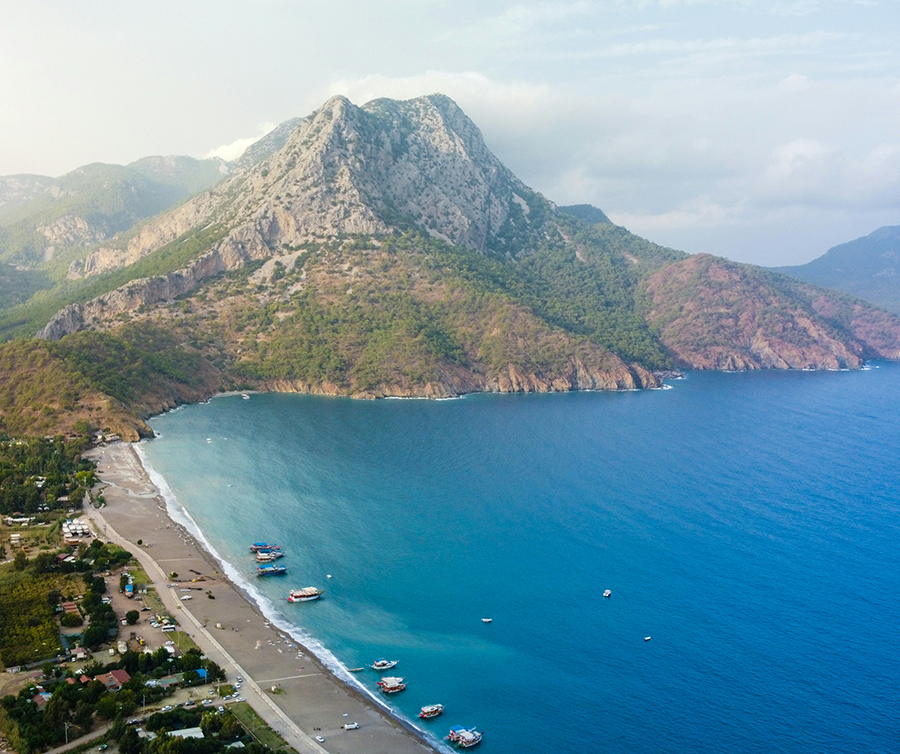 Antalya gezi: Antalya'nın Kumluca ilçesine bağlı Adrasan Koyu'nu kuşbakışı gösteren bu fotoğrafta, turkuaz renkli denizin yemyeşil dağlarla buluştuğu uzun bir sahil şeridi, kıyıya yanaşmış tekneler ve sahildeki yapılaşma doğal güzelliğin bir parçası olarak görülüyor.