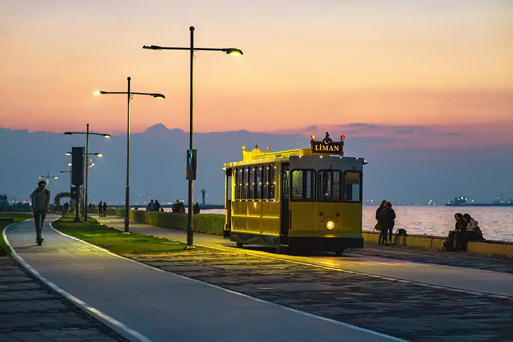 Gün batımında Alsancak Kordonu'nda, denizin kenarında ilerleyen sarı bir nostaljik tramvay ve ışıklı sokak lambaları eşliğinde yürüyüş yapan insanlar, arka planda belirginleşen dağ silüetleriyle huzurlu bir şehir manzarasını yansıtıyor.