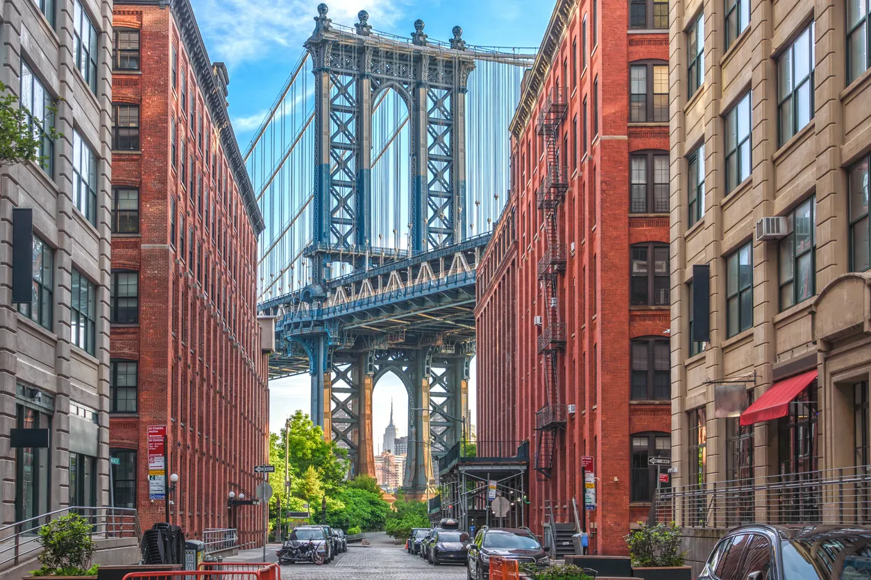 Amerika'daki bahar rotaları için harika bir durak olan New York'un ikonik Manhattan Köprüsü'nün, kırmızı tuğlalı ve taş binalar arasında belirgin bir şekilde göründüğü ve uzakta bir gökdelenin seçilebildiği, ağaçların yeşillendiği, park etmiş araçlarla dolu pitoresk bir sokak manzarası.