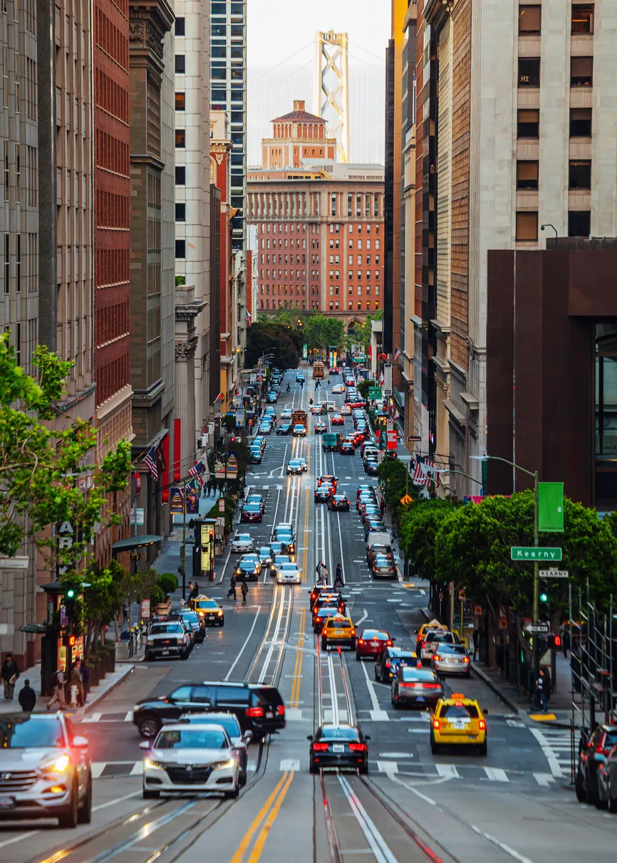 San Francisco'nun simgesel dik yokuşlarından aşağı uzanan, araçlarla yoğun Kearny Caddesi'nde kalabalık şehir hayatı ve yeşil ağaçlar görülüyor; uzakta ise Bay Köprüsü'nün zarif yapısı ve tarihi tuğla binalar Amerika bahar rotası deneyimine eşlik ediyor.