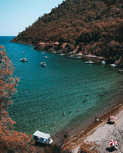 Antalya'nın kızılımsı ağaçlık yamaçların kucakladığı berrak turkuaz sulara sahip bir koyunda, yüzen tatilciler, demirlemiş tekneler ve sahile yakın konumlanmış bir kamp aracı, Akdeniz'in eşsiz doğal güzelliklerini ve dingin bir seyahat atmosferini gözler önüne seriyor.