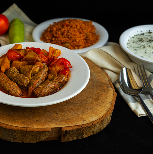 Ahşap servis tahtası üzerinde, Balkan Lokantası'nın öne çıkan esnaf lezzetlerinden biberli köfte, nar gibi kızarmış bulgur pilavı ve yanında ferahlatıcı cacık içeren geleneksel bir sofra düzeni, Sinem Özler'in ve diğer şeflerin favori lezzet duraklarını keşfetmeye davet ediyor.