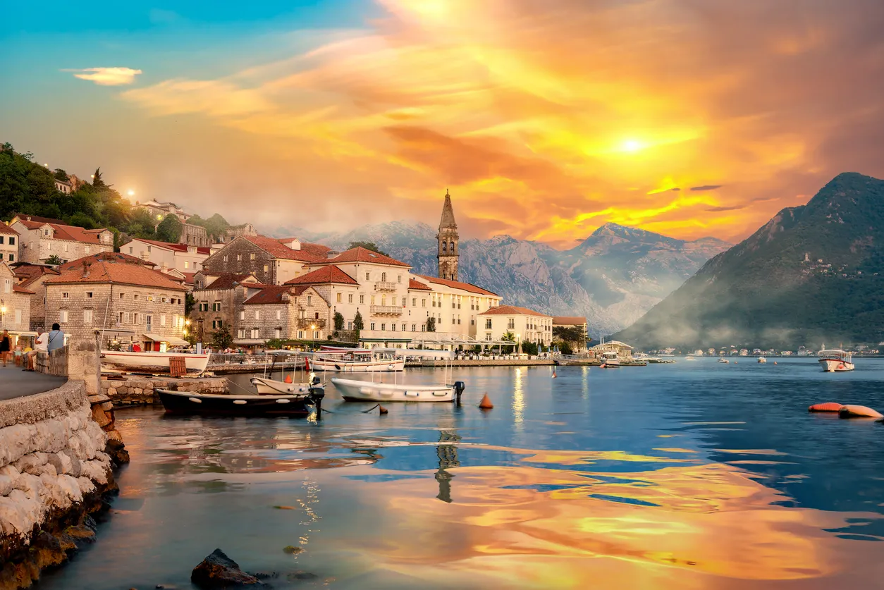 Balkanlar'daki Perast benzeri tarihi bir sahil kasabasında, dağların çevrelediği koyun üzerinde batan güneşin turuncu ve sarı tonlarıyla parlayan gökyüzünün suya yansıdığı, geleneksel taş binaların ve kıyıya demirli teknelerin büyüleyici bir bahar rotası manzarasını sergileyen geniş bir görsel.
