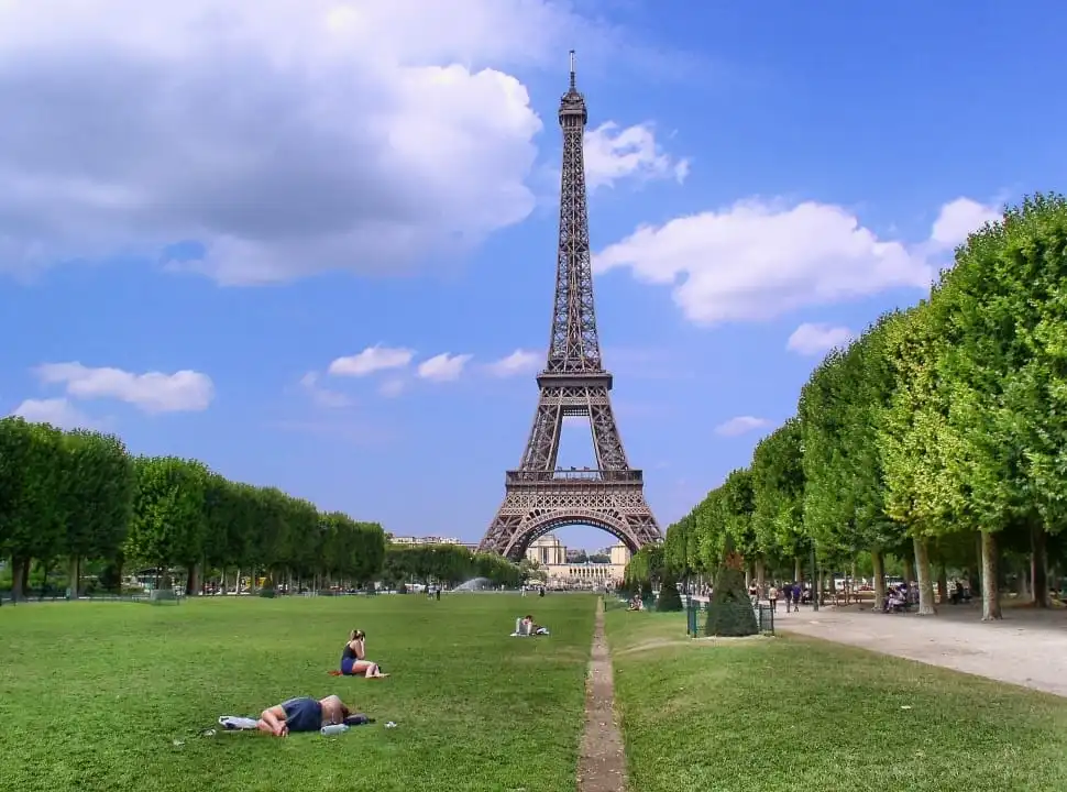 Eyfel Kulesi'nin heybetli silüeti altında, Paris'in yemyeşil Champ de Mars parkında güneşli bir günde çimlere uzanmış ve dinlenen insanlar, ziyaretçiler için huzurlu bir Paris Gezi Rehberi sahnesi sunuyor.