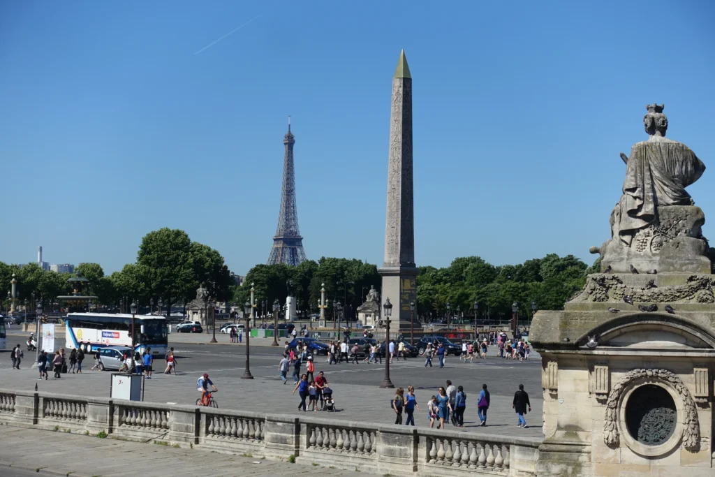 Paris'teki Concorde Meydanı'nda, güneşli ve açık mavi gökyüzü altında, sağda bir heykelin ve önde taş korkuluğun görüldüğü, Luxor Dikilitaşı ile uzaktaki Eyfel Kulesi'nin yükseldiği, kalabalık bir şehir manzarası.