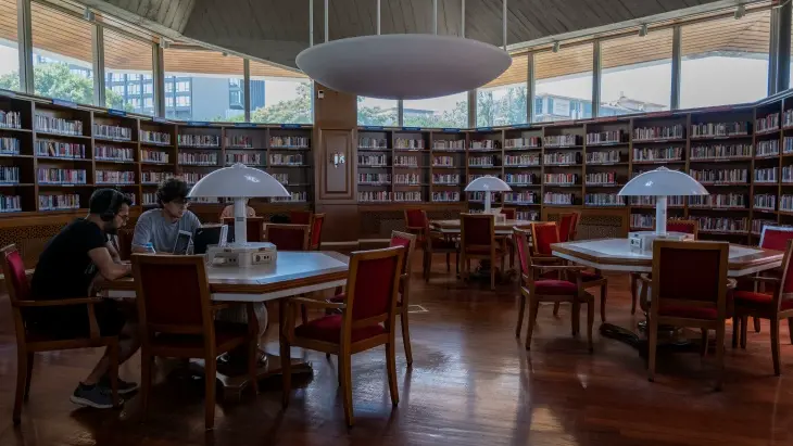 İstanbul'un en iyi kütüphanelerinden biri olan Atatürk Kitaplığı'nda, dairesel ahşap raflarla çevrili, yüksek pencerelerden doğal ışık alan geniş bir salonda, iki kişinin laptoplarıyla çalıştığı, mantar şekilli lambalarla aydınlatılmış beyaz masalar ve kırmızı sandalyeler, zamanın yavaşladığı bir çalışma ve okuma atmosferi sunuyor.