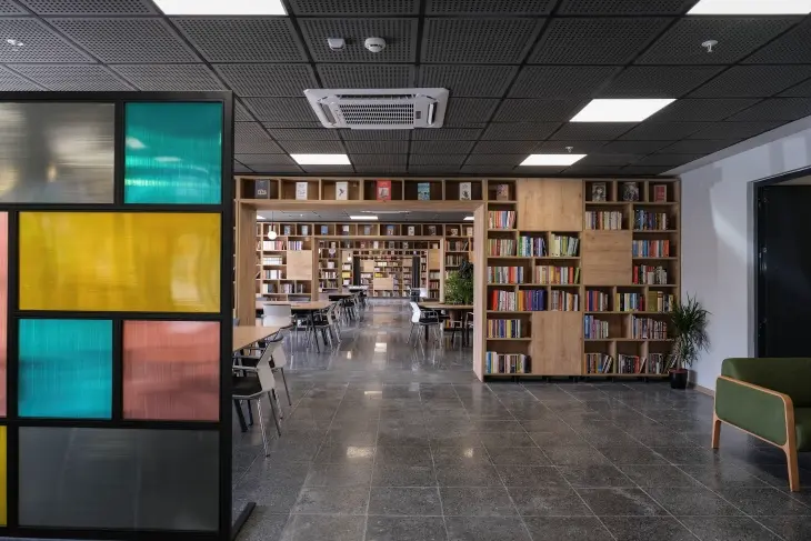 Modern tasarımlı, renkli cam panelli bir bölücü önünde, ahşap raflara düzenli bir şekilde dizilmiş yüzlerce kitabın bulunduğu geniş ve aydınlık bir kütüphane iç mekanında, gri karo zemin ve delikli tavan panelleri dikkat çekiyor; bu yeni nesil kütüphane, İstanbul’un en iyi okuma ve çalışma alanlarından birini sunuyor.