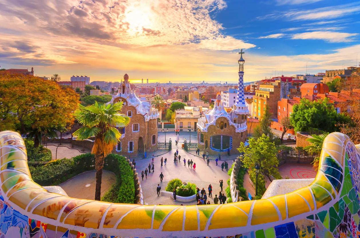 İspanya bahar rotasının gözde duraklarından Park Güell'de, rengarenk mozaiklerle bezeli banklardan görünen, Gaudi'nin kendine özgü mimarisiyle inşa edilmiş iki masalsı yapı ve kalabalık ziyaretçilerle dolu meydan, arkada gün batımının kızıla boyadığı Barselona şehir silüetiyle bütünleşiyor.