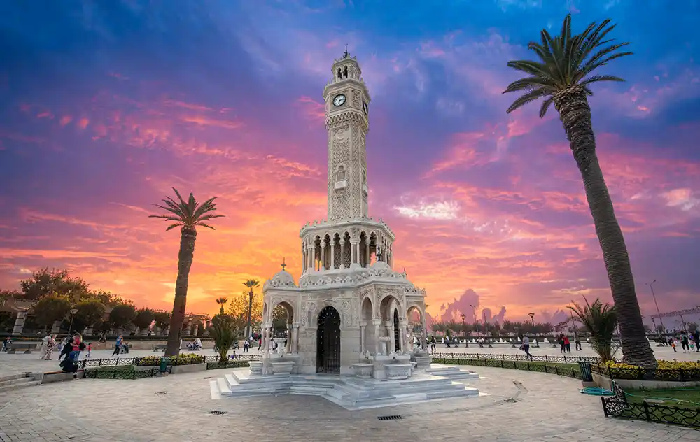 İzmir Saat Kulesi, Konak Meydanı'nda göz alıcı turuncu, pembe ve mor renklerdeki dramatik gün batımı gökyüzünün altında, çevresindeki palmiye ağaçları ve yeşilliklerle birlikte zarif mermer mimarisi ve dört bir yanındaki saatleriyle ikonik bir sembol olarak yükseliyor.