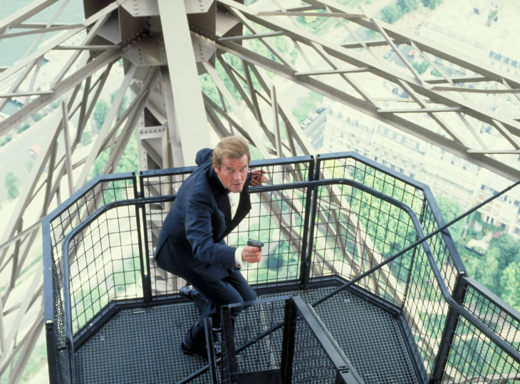 En İyi James Bond Filmleri serisinden Roger Moore, ikonik Eyfel Kulesi'nin yüksek metal platformunda, elinde küçük bir tabanca tutarak tetikte ve kararlı bir şekilde eğilmiş halde, arkasında Paris şehrinin geniş bir manzarası uzanıyor.