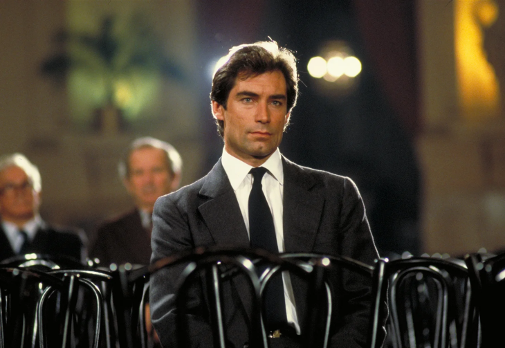 James Bond filmleri serisinde karaktere ağırlıklı bir duruş kazandıran Timothy Dalton, koyu gri takım elbisesi, beyaz gömleği ve siyah kravatıyla ciddi bir ifadeyle kameraya bakarken, arkasında flu görünen diğer katılımcılarla birlikte resmi bir toplantı veya dinleme salonunda sandalyelerin önünde oturuyor.