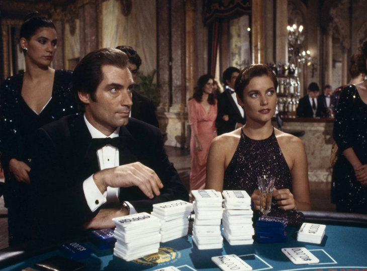 Takım elbiseli Timothy Dalton'ın canlandırdığı James Bond ve kısa saçlı Maryam d'Abo, lüks bir kumarhanede masada otururken, önlerindeki oyun fişleri James Bond filmlerinin ikonik casusluk ve gerilim dolu atmosferini yansıtan sahnenin heyecanını artırıyor.