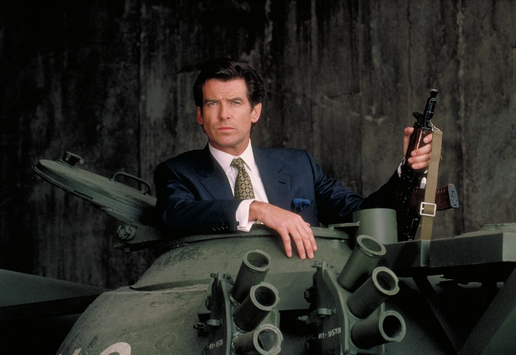 Pierce Brosnan, koyu renk takım elbisesiyle bir tankın açık kapağından dışarı doğru bakarken, elinde bir tüfek tutarak objektife kararlı bir ifadeyle bakıyor ve En İyi James Bond Filmleri serisine damga vuran aksiyon sahnelerinden birini çağrıştırıyor.