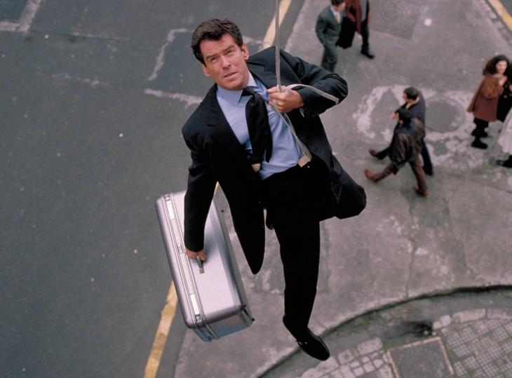 James Bond filmlerinden bir sahneyi yansıtan karede, Pierce Brosnan, şehir sokağının üzerinde bir halatla asılı dururken, sol elinde gümüş renkli bir evrak çantası taşıyor; kararlı ifadesi ve aşağıdaki kalabalığın farkında olmaması, casus karakterinin tehlikeli dünyasını gözler önüne seriyor.