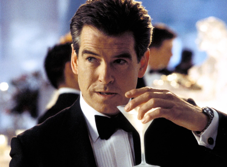 Siyah smokini, beyaz pilili gömleği ve papyonuyla şık bir görünüm sergileyen Pierce Brosnan, James Bond karakteriyle elinde ikonik martini kadehini tutarken, karizmatik bakışıyla casus filmleri efsanesine damga vuran En İyi James Bond Filmleri dönemini yansıtıyor.