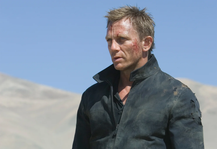 Daniel Craig, yüzündeki kan ve kir lekeleriyle, yıpranmış ceketinin içinde çöle benzer bir manzara önünde ciddi bir ifadeyle James Bond olarak dururken, En İyi James Bond Filmleri serisinin aksiyon dolu ve daha gerçekçi anlatımını yansıtıyor.