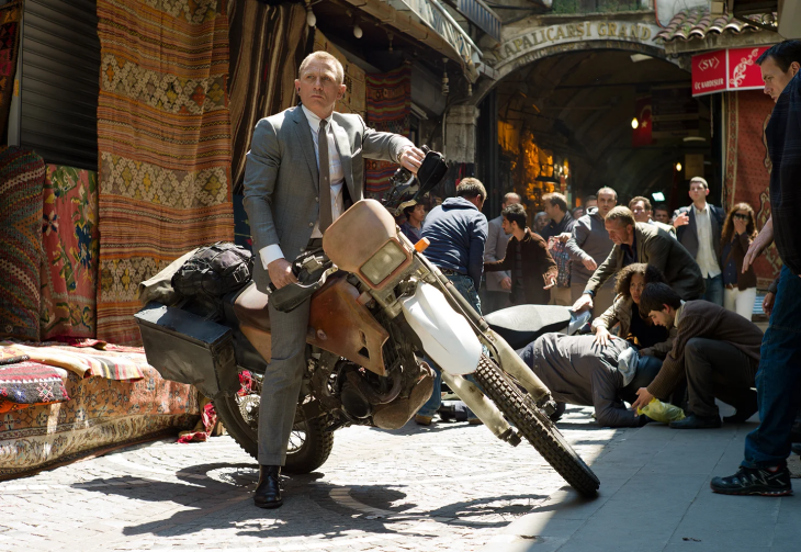 Daniel Craig, gri takım elbisesiyle İstanbul Kapalıçarşı'da bir motosikletin üzerinde dururken, etrafındaki kalabalık bir olaya müdahale ediyor ve bu an En İyi James Bond filmlerinden Skyfall'un aksiyon dolu sahnelerinden birini yansıtıyor.