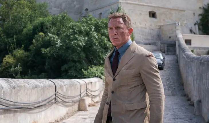 James Bond Filmleri serisinden Daniel Craig, yüzünde hafif kir izleriyle ve kararlı, ciddi bir ifadeyle, kahverengi bir ceket ve lacivert bir gömlek giymiş, tarihi taş bir köprüde ilerlerken, arkasında yeşil ağaçlar ve antik bir kale yapısı yükseliyor.