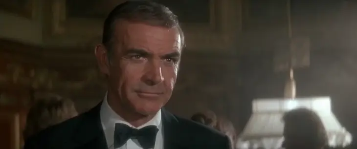 En iyi James Bond filmleri listemizin efsanevi yüzlerinden Sean Connery, karanlık ve şık bir iç mekanda siyah smokini ve papyonuyla doğrudan izleyiciye bakarken, casus karakterinin karizmatik ve kararlı duruşunu sergiliyor.
