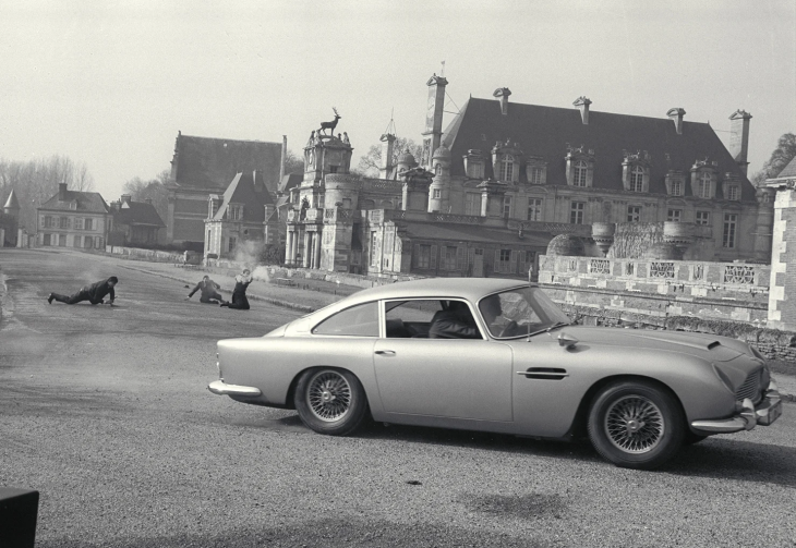 Gri renkli, klasik bir Aston Martin DB5 araba, sağ ön planda dururken, sol arka planda büyük bir şatonun önündeki yolda üç kişi bir aksiyondan kaçınır gibi yerde sürünüyor, ikonik James Bond filmlerinin atmosferini yansıtıyor.