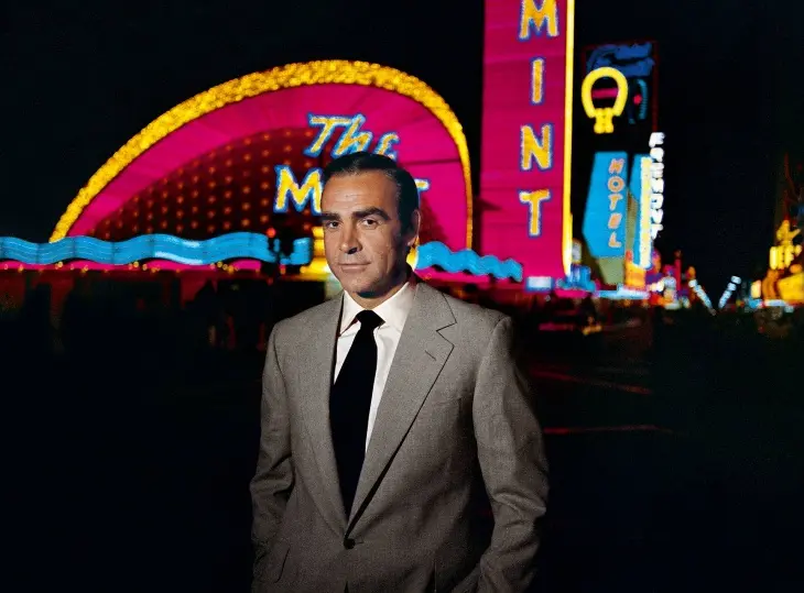 En İyi James Bond Filmleri serisinin efsanevi aktörü Sean Connery, geceleyin parlak pembe, sarı ve mavi neon ışıklarla aydınlatılmış The Mint gibi bir kumarhane veya otelin önünde, karizmatik bir duruşla izleyiciye bakıyor.