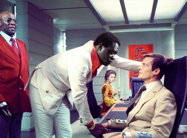 Live and Let Die filminden Roger Moore'un canlandırdığı James Bond, kolu metal bir aparatla sabitlenmiş halde sandalyede otururken, beyaz takım elbiseli iri bir adam tarafından sorgulanmakta, arkada kırmızı takım elbiseli ikonik düşman Baron Samedi ve altın rengi elbiseli bir kadın bu gergin casusluk sahnesini dikkatle izlemektedir.