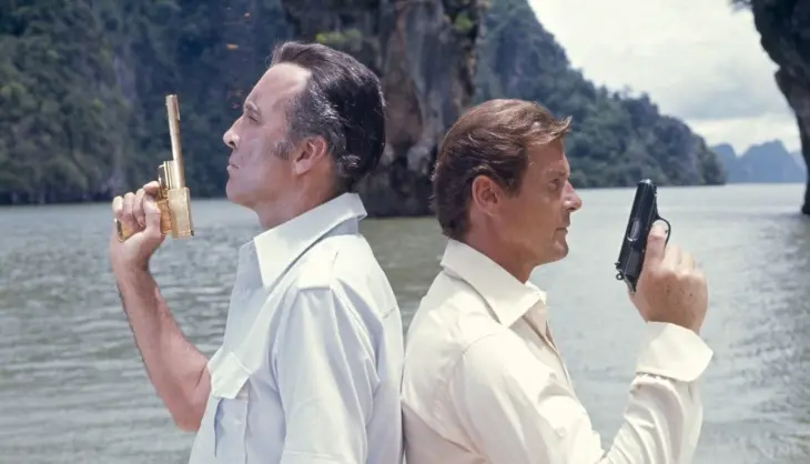 Roger Moore'un canlandırdığı James Bond, klasik siyah tabancasıyla, arka planda yemyeşil tropik adaların yükseldiği bir su yolunda, sırtı dönük vaziyette, efsanevi suikastçı Scaramanga'nın altın kaplamalı silahını tutarken gergin bir anı gösteriyor; bu sahne En İyi James Bond Filmleri listesindeki önemli yapımlardan biri olan Altın Tabancalı Adam filminden bir kesit.