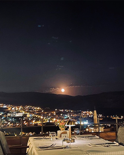 Kapadokya'nın ışıltılı şehir manzarasının ve tepelerin üzerinde parlayan dolunayın eşlik ettiği, beyaz örtülü bir restoran masasının gece çekimi, bölgedeki en iyi mekanlar arasında unutulmaz bir gastronomi deneyimi vadediyor.