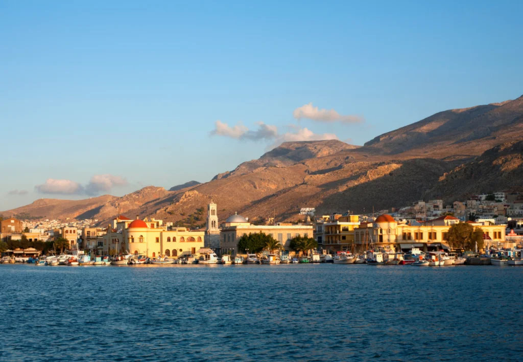 Yunanistan'ın Kalymnos adasında, parlak turkuaz denizin kıyısında sıralanmış, kırmızı kiremitli çatılara ve pastel tonlara sahip geleneksel binaların, bir kilise kulesinin ve limanda demirli teknelerin oluşturduğu, arka planda heybetli dağların yükseldiği güneşli bir sahil kasabası manzarası.