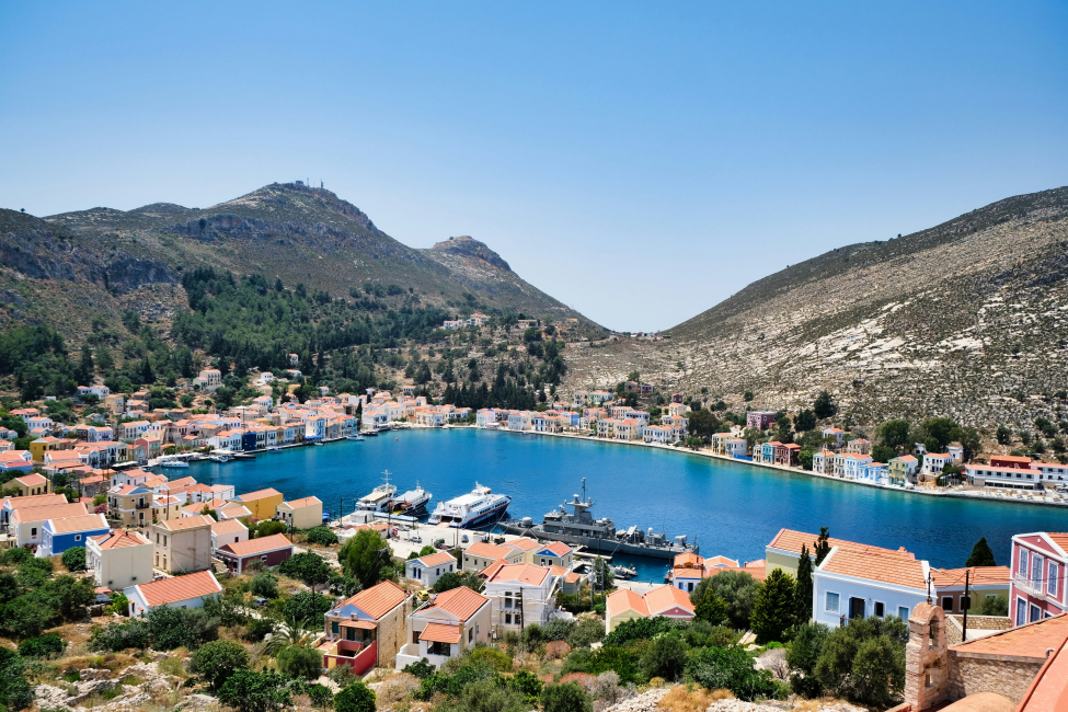 Yunanistan'ın pitoresk Kastellorizo Adası'nda, canlı renkli evlerin masmavi bir koyu sardığı, dik yamaçlarda yükselen yeşilliklerin ve berrak gökyüzünün eşlik ettiği, teknelerin demirli olduğu huzurlu bir liman manzarası.