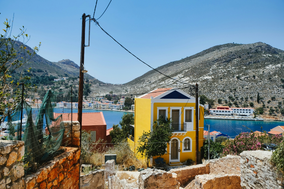 Kastellorizo'nun canlı ruhunu yansıtan bu manzarada, parlak sarı renkli, mavi alınlıklı ve iki katlı bir ev ön planda yer alırken, arkasında masmavi bir koy, renkli binalar ve Ege Denizi'nin kurak dağlarla buluştuğu eşsiz Yunanistan coğrafyası uzanıyor.