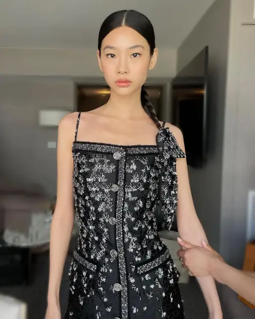 Model, ortadan ayrılmış, geriye doğru toplanmış örgülü at kuyruğu saç modeliyle doğrudan kameraya bakıyor; at kuyruğunun dibine bağlı siyah bir kurdele saç aksesuarı olarak şık ve zarif bir dokunuş katarken, üzerinde beyaz işlemeler olan askılı siyah elbisesiyle kurdeleyle yapabileceğiniz saç modelleri arasında trend bir örnek sunuyor.