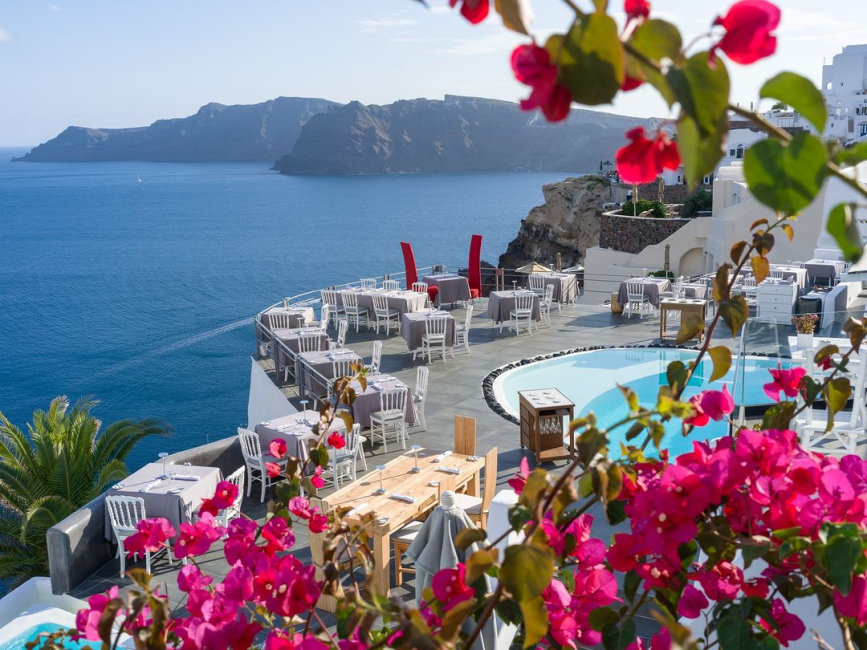 Santorini'nin volkanik manzarası eşliğinde, pembe begonvillerle çerçevelenmiş bu açık hava restoranı, Ege Denizi'nin derin mavisini kucaklayan terasında şık gri örtülü masaları ve rahatlatıcı bir havuz alanıyla Yunan Adaları'nda unutulmaz bir yemek deneyimi sunuyor.