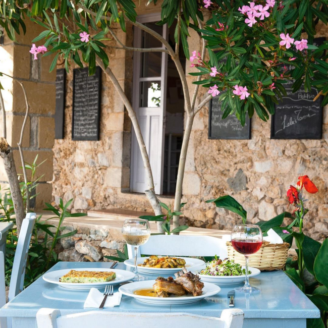 Lela's Taverna'nın taş cephesinin önünde, pembe zakkumların gölgesindeki açık mavi ahşap masada, kuzu pirzola, fırın yemeği, taze salata ve iki kadeh şarapla dolu, Yunanistan restoranlarının samimi atmosferini yansıtan keyifli bir açık hava öğle yemeği.