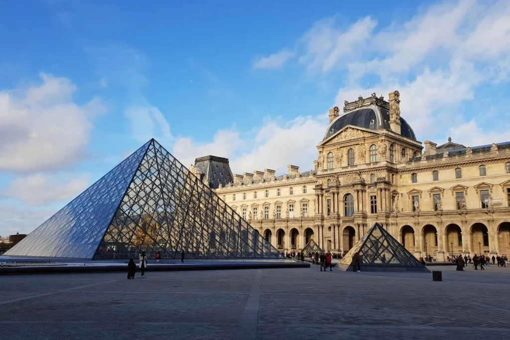 Masmavi ve bulutlu bir gökyüzünün altında, geniş bir meydanda yer alan ikonik Louvre Müzesi'nin büyük cam piramidi ve arkasındaki Barok tarzı görkemli saray binası, etrafta dolaşan ziyaretçilerle birlikte göz alıcı bir manzara sunuyor.