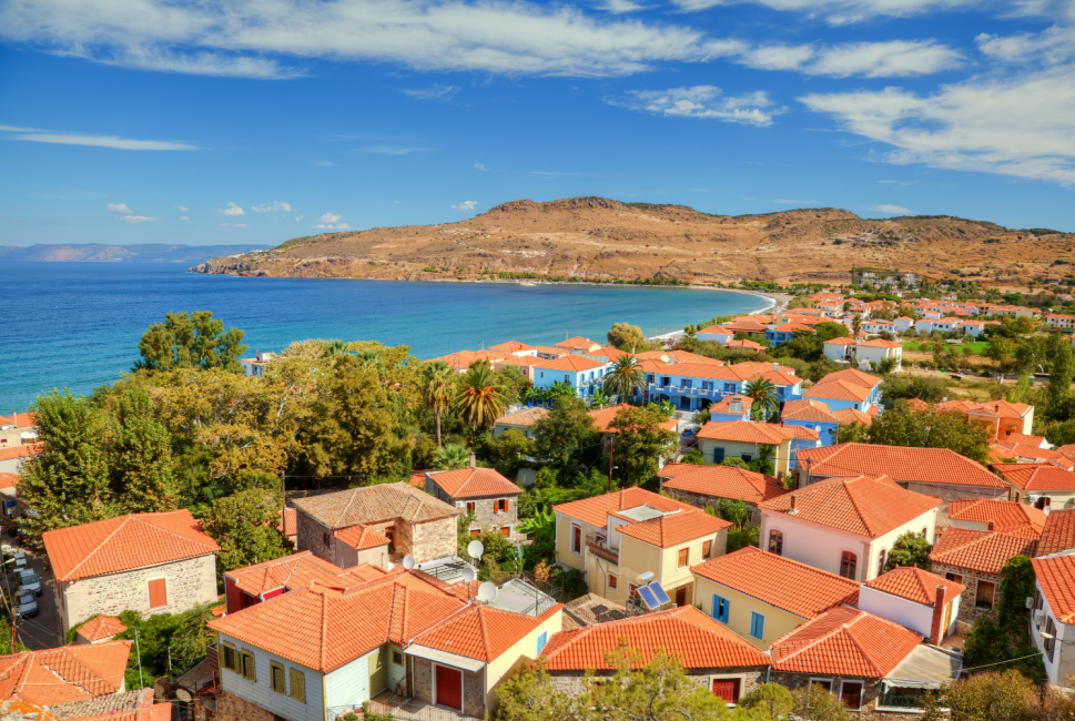 Midilli Adası'nda, Kapıda Vizeyle gidilebilecek popüler Yunan Adaları'ndan biri olan sahil kasabasının kuşbakışı görünümü, canlı turuncu çatılı evleri, palmiye ağaçları, berrak turkuaz denizi ve arkadaki kahverengi tepelerle güneşli bir yaz gününü gözler önüne seriyor.