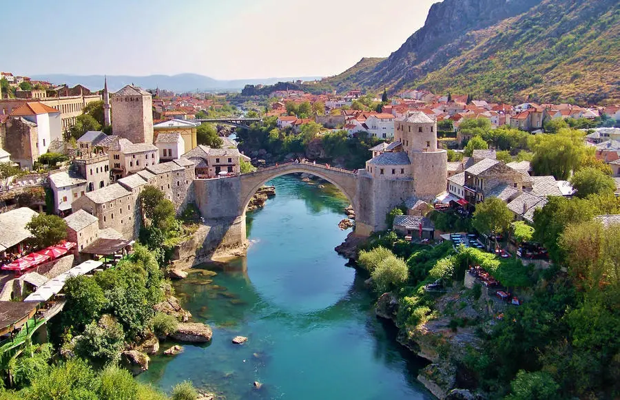 Mostar Köprüsü'nün ikonik kemerli yapısı, berrak turkuaz rengi Neretva Nehri üzerinde yükselirken, her iki kıyıda da tarihi taş yapılar, yemyeşil ağaçlar ve arka planda uzanan dağlarla çevrili pitoresk bir şehir manzarası sunuyor.