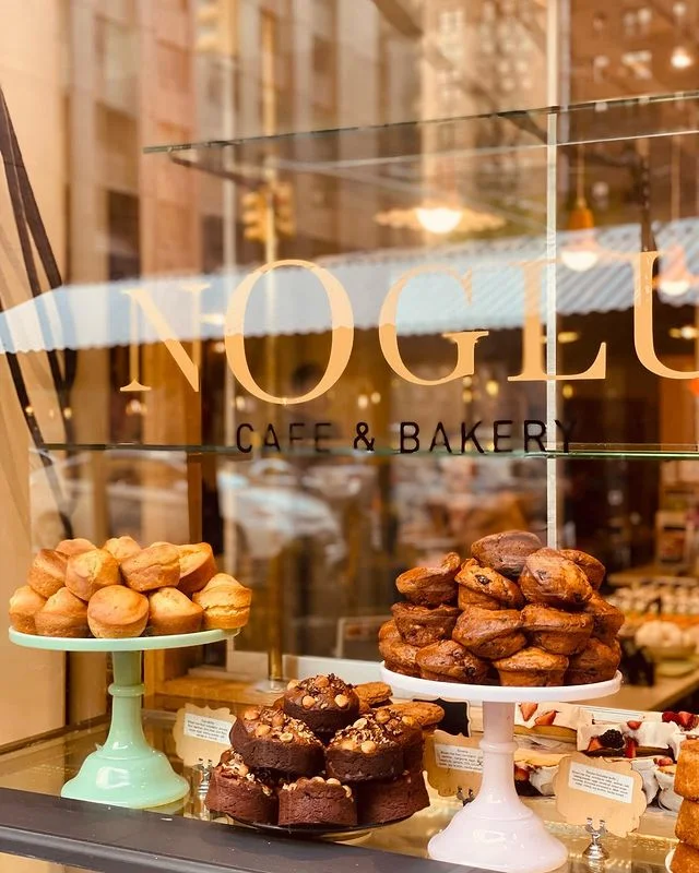 Noglu Cafe & Bakery'nin vitrininde, açık yeşil bir stand üzerinde bolca sade muffin, ortada fındık ve ceviz parçacıklarıyla süslenmiş yoğun çikolatalı küçük kekler ve pembe bir standda çeşitli meyveli muffinler iştah açıcı bir şekilde sergileniyor.