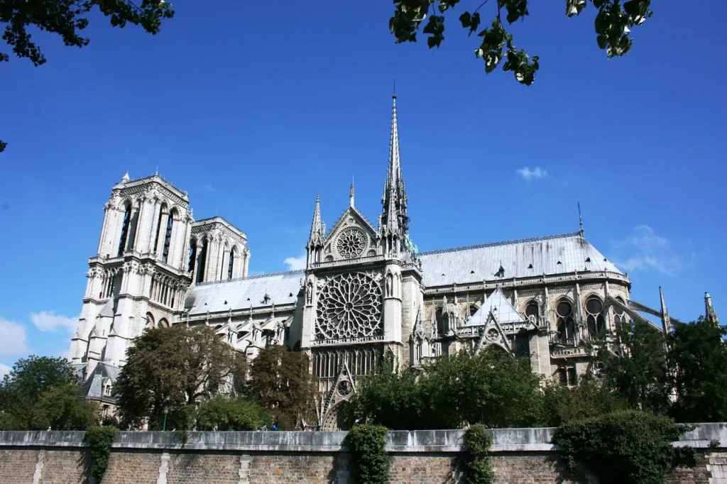 Güneşli parlak bir gökyüzünün altında yükselen Paris'in ikonik Notre Dame Katedrali, karakteristik ikiz çan kuleleri, büyük merkezi gül penceresi ve sivri kulesiyle görkemli Gotik mimarisini sergilerken, ön planda Seine Nehri kıyısındaki taş duvar ve yemyeşil ağaçlar yer almaktadır.