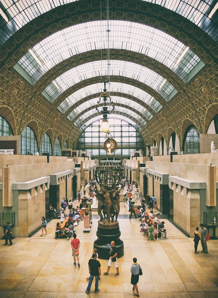 Orsay Müzesi'nin geniş, kemerli, cam tavanlı ana salonunda, ortada dünya figürünü taşıyan üç bronz heykel bulunuyor ve ziyaretçiler tarihi atmosferde dolaşarak sanat eserlerini inceliyor.