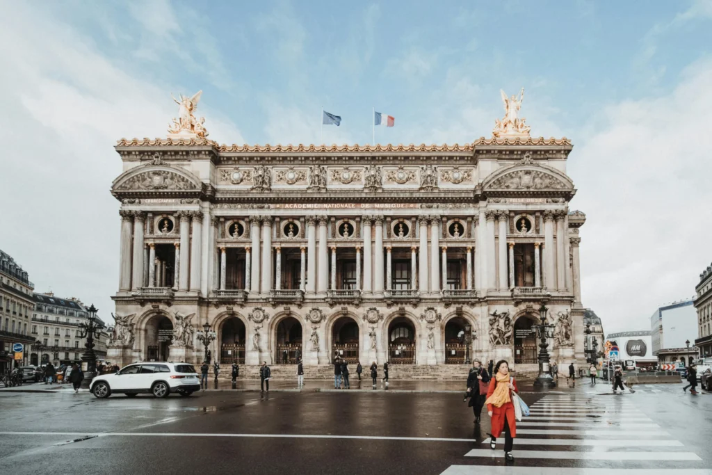 Paris Gezi Rehberi'nin mimari güzelliklerine ışık tutan bu karede, Palais Garnier'nin altın detayları, heykelleri ve bayraklarıyla süslü görkemli cephesi, ıslak zeminde yürüyen insanlarla adeta bir film sahnesini andırıyor.
