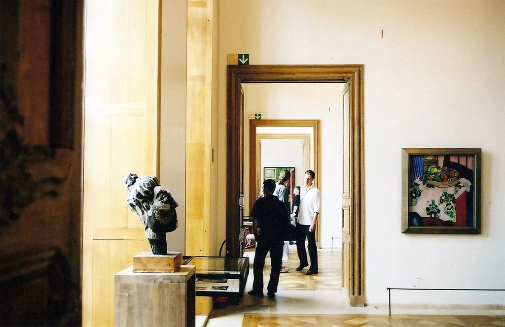 Aydınlık bir modern sanat müzesi salonunda, ahşap bir podyumda sergilenen dokulu soyut bir büst heykeli ile duvarda asılı canlı renklerde meyve sepeti natürmort tablosu göze çarparken, ziyaretçiler art arda sıralanmış odalara açılan kapılardan geçiyor.