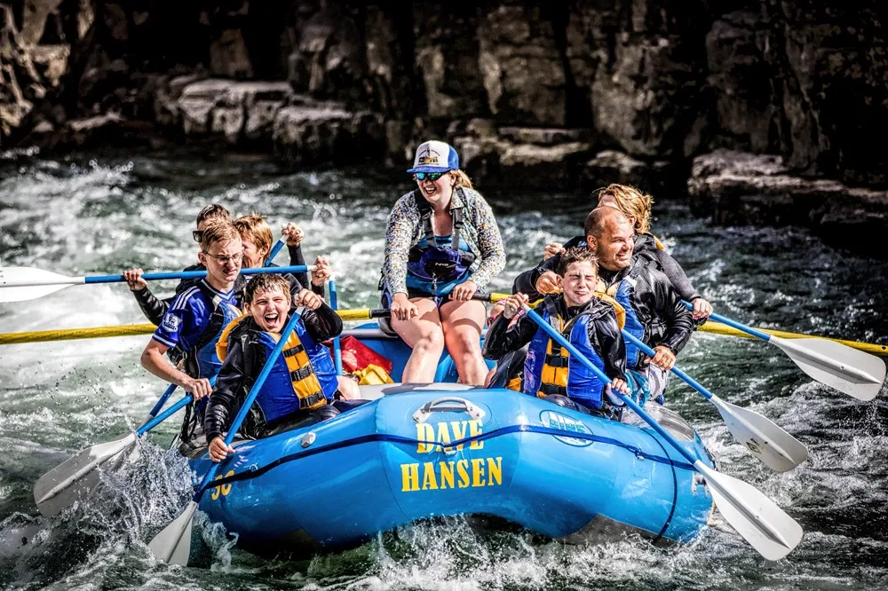 Bir grup yetişkin ve çocuk, can yelekleri giyerek Dave Hansen yazılı mavi bir şişme botta coşkulu bir şekilde beyaz sularda rafting yaparken kürek çekiyor; nehrin kayalık kıyıları arka planda kalırken, ekip üyelerinin yüzlerindeki heyecan ve mutluluk dalgalarla birlikte yansıyor.