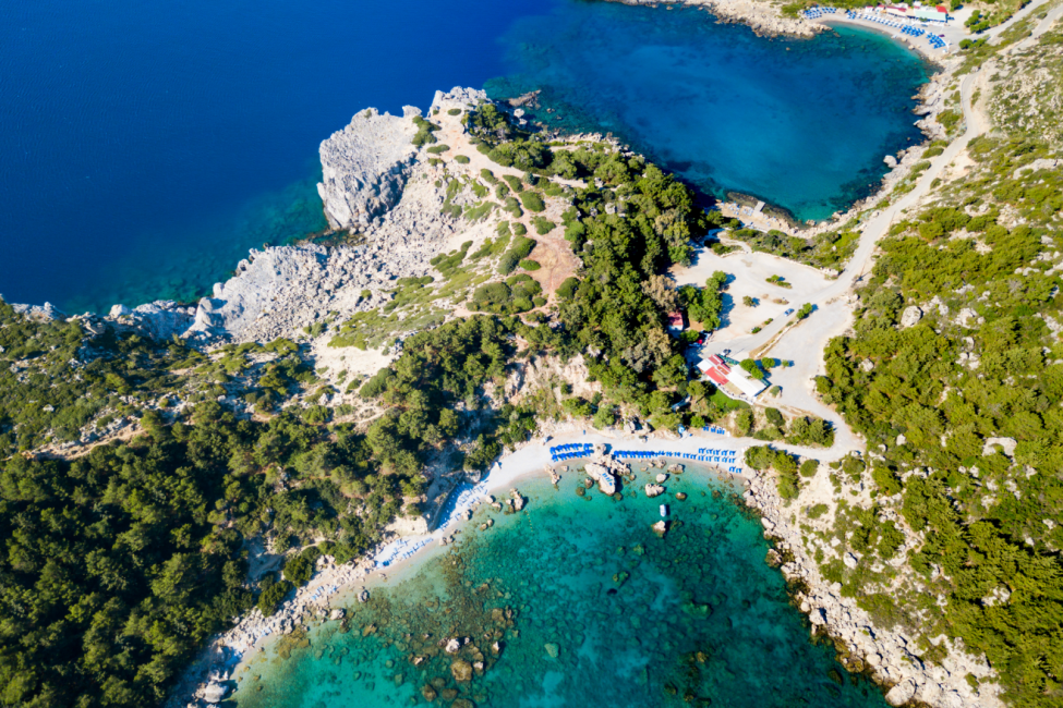 Rodos Adası'nın kapıda vize uygulamasıyla ziyaret edilebilecek cazip Yunan Adaları'ndan biri olduğunu vurgulayan bu kuşbakışı görüntüde, yemyeşil tepelerle çevrili berrak turkuaz sulara sahip bir koyda uzanan şezlong ve şemsiyelerle donatılmış kumlu bir plaj ile çevresindeki kayalık kıyı şeridi ve masmavi deniz göz alıyor.