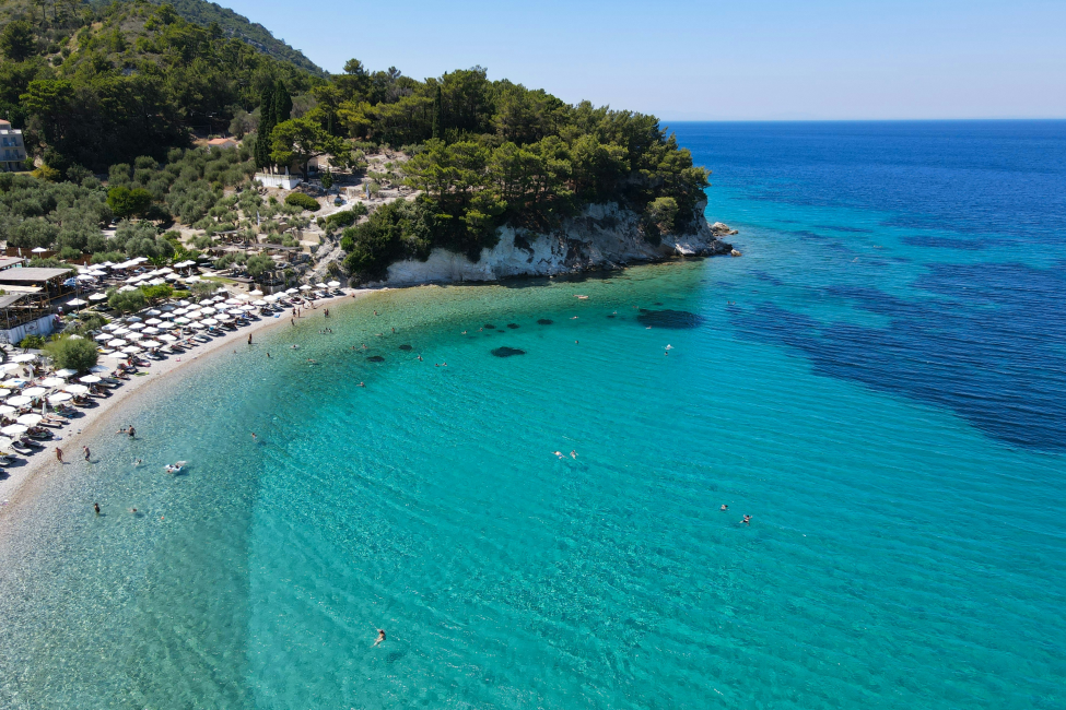 Yunanistan'ın Samos Adası'nda, bembeyaz şemsiyelerle dolu kalabalık bir çakıllı plaj, turkuazdan laciverte dönen berrak denizi ve yemyeşil tepelerin kuşattığı bu doğal güzelliği kuşbakışı sergiliyor.
