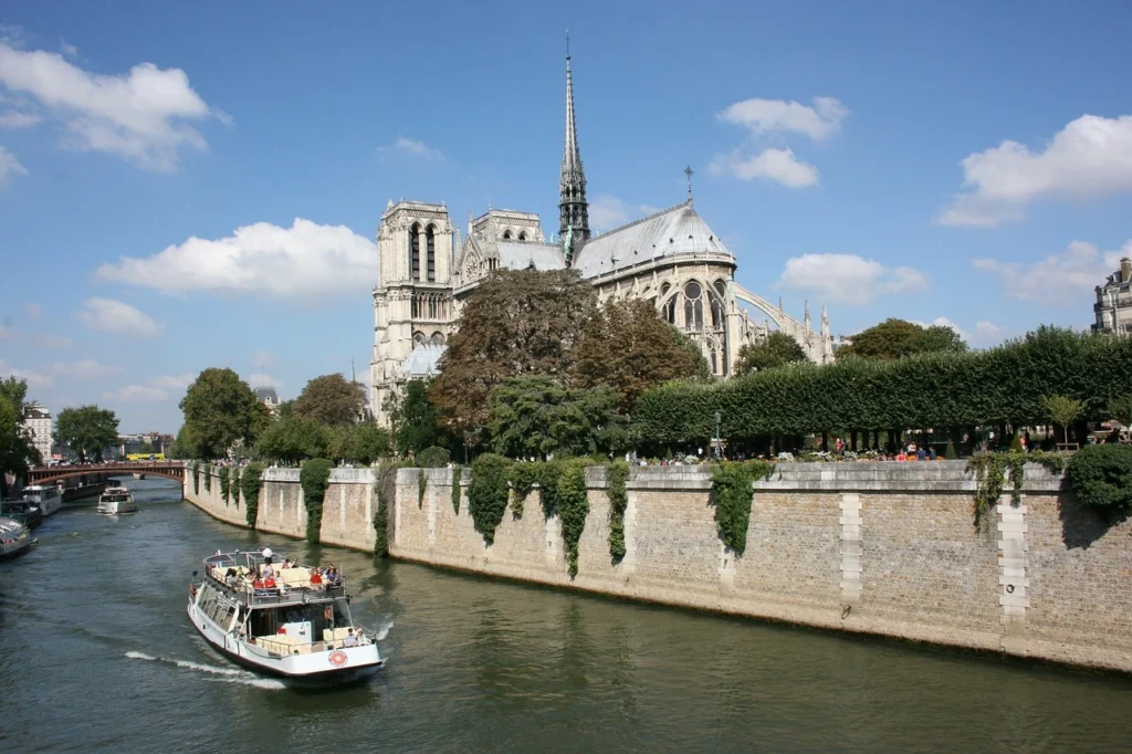 Paris'teki Notre Dame Katedrali'nin kuleleri ve sivri çatısıyla birlikte, üzerinde bir gezi teknesinin ilerlediği Seine Nehri'nin taş duvarlı ve yeşilliklerle kaplı kıyıları güneşli bir havada gökyüzündeki bulutların altında görülüyor.