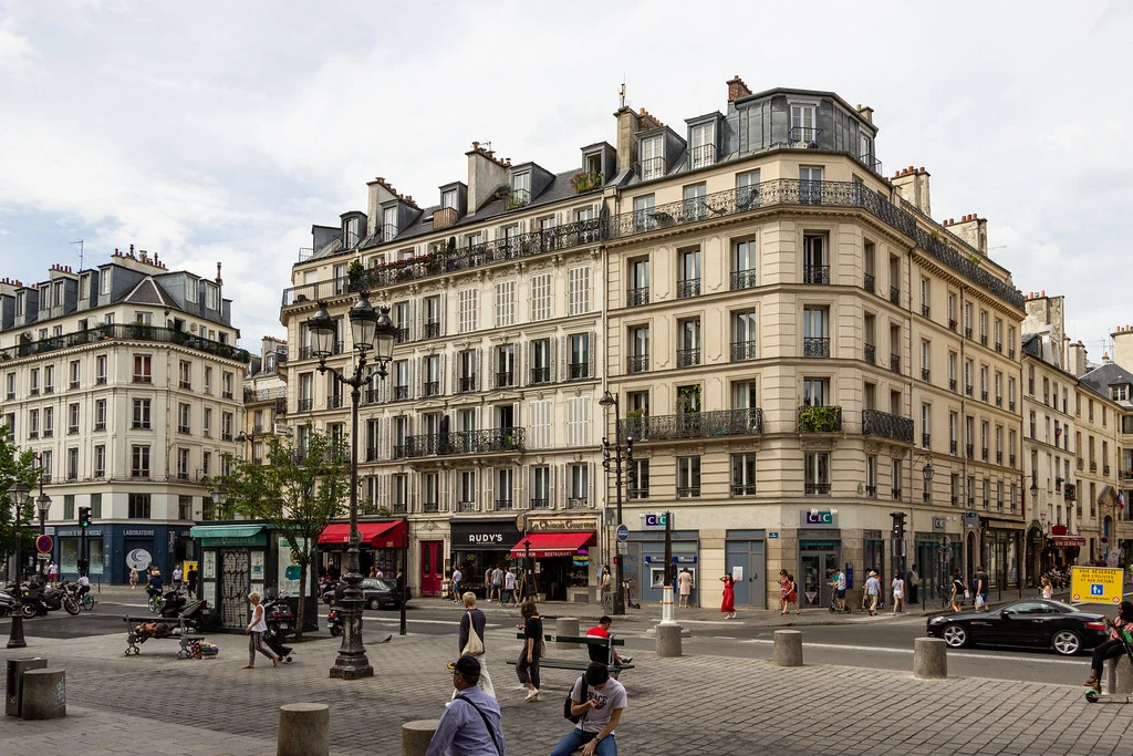 Haussmann tarzı, çok sayıda pencere ve demir balkonlara sahip binaların sıralandığı, zemin katlarında mağazaların ve bir bankanın bulunduğu hareketli bir Paris alışveriş caddesi görülen bu geniş açılı görselde, kaldırımlarda dolaşan birçok insan ve caddede ilerleyen siyah bir otomobil dikkat çekmektedir.
