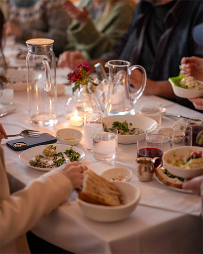 Ayvalık'ın eşsiz gastronomik zenginliğini yansıtan, beyaz masa örtüsü üzerinde mum ışıkları ve çiçeklerle süslü bir akşam yemeği sofrasında, çeşitli mezeler, taze ekmekler ve içeceklerle çevrili insanların Kuzey Ege'nin tadını çıkararak keyifli anlar paylaştığı an.