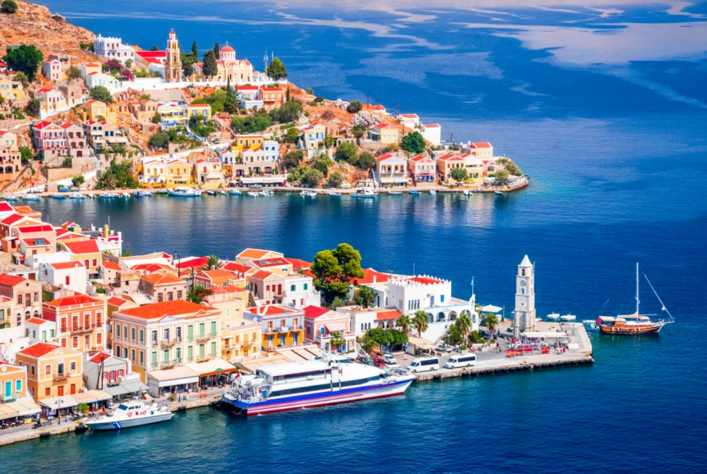 Yunanistan'ın Symi Adası'nda, tepelere kurulu rengarenk evlerin masmavi denize baktığı, limanda teknelerin demirli olduğu ve sahilde beyaz bir saat kulesinin yükseldiği büyüleyici bir kıyı kasabası manzarası.